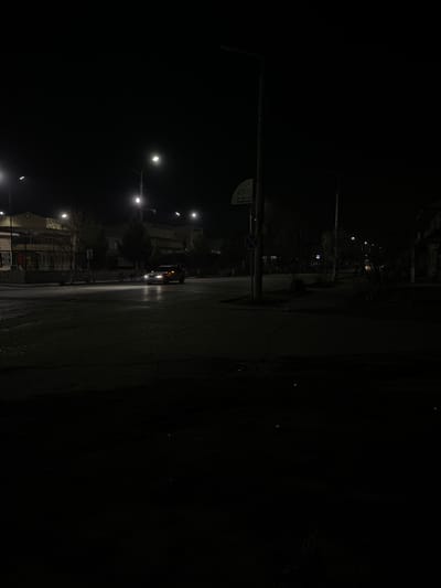 The image displays a nighttime street scene in Karmana, Uzbekistan. The asphalt road surface is visibly wet and reflective from artificial light sources. A light-colored sedan, with its headlights activated, is observed in motion across the mid-ground, traveling from left to right. No human subjects are discernible within the frame. On the left side, multi-story buildings with minimal interior illumination are partially visible behind a dark fence. The right side of the street features darker, less defined structures receding into the background. Multiple utility poles support streetlights, casting distinct pools of light. One prominent utility pole in the central-right foreground exhibits a circular blue sign with a red diagonal line, indicating a 'no stopping' or 'no parking' restriction, and an indecipherable white rectangular sign mounted above it. Sparse, dormant trees and shrubs line the pavement edges. The environment appears to be an urban setting, devoid of activity beyond the moving vehicle.