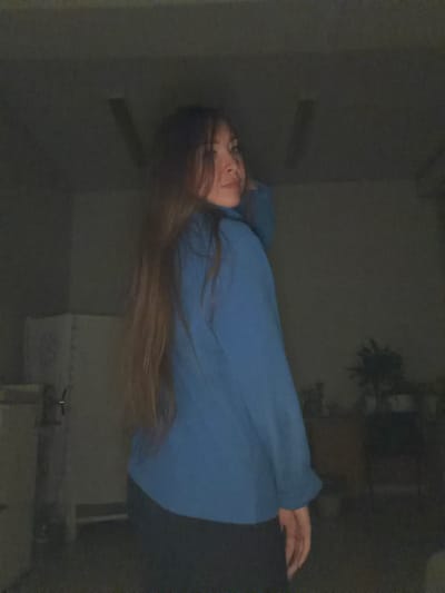 In a dimly lit interior space, a woman with long, dark brown hair is captured from the mid-torso up, posing with her back partially turned to the camera. She is wearing a blue long-sleeved shirt and black pants. Her head is turned to her right, and she is looking over her shoulder towards the camera, with her left hand resting on her hip. The background is dark and out of focus, with some indistinct objects and furniture visible. There is a faint light source illuminating the scene from the front. The environment appears to be an indoor location, possibly a room or a studio, with a concrete floor. The overall impression is that of a candid or posed photograph taken in low-light conditions. There is no information available that would indicate this scene is located in the city of Япрыково, Russia.