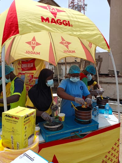 This media file captures an outdoor promotional event for Maggi products in Tarauni, Nigeria. The scene is set under a large yellow and red Maggi-branded tent. Several individuals, likely employees or volunteers, are actively participating. Two people in the foreground, wearing blue hairnets and face masks, are preparing food. One person, wearing a blue t-shirt with "PL PARADOL" printed on it, is adding a packet of Maggi seasoning into a pot on a portable gas stove. Another person, partially visible in a yellow shirt and black hijab, is also wearing a face mask and seems to be assisting with the cooking. To the left, another individual in a yellow shirt and blue hairnet is also wearing a face mask.

Several objects are prominent in the scene. To the left, a yellow box labeled "MeWe Blender & Grinder" is visible, along with other Maggi product packaging in the background. Water bottles, cups, and cooking utensils are arranged on a white table in front of the food preparation area. A blue gas stove is the central focus of the cooking activity. The overall atmosphere appears to be a promotional setup, possibly a cooking demonstration or sampling event. The bright sunlight suggests it is daytime, and the presence of the tent indicates an outdoor event. The background shows a slightly cluttered environment with some construction materials, suggesting a less developed area or a street-side location. The text visible includes "MAGGI", "CHOOSE THE STAR", "MeWe Blender & Grinder", and various product specifications in English, French, and Portuguese.