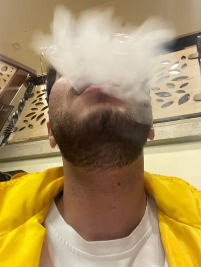 The image displays an upward-angled view of an adult male within an indoor establishment, identified as being in Homs, Syria. The subject, who possesses a trimmed beard on his chin and jawline, is observed exhaling a substantial volume of dense, white vapor from his mouth, which largely obscures his upper facial features. A clear, rounded object, consistent with a shisha mouthpiece, is partially discernible within the vapor plume directly in front of his mouth. He is attired in a white crew-neck t-shirt underneath a bright yellow collared jacket. The background reveals interior architectural elements: a light-colored wall on the right featuring a decorative wooden panel with multiple irregular, elongated cut-out patterns, and a darker wooden structure on the left with turned vertical elements and carved details, indicative of a partition or railing. A square recessed light fixture is visible on the light-colored ceiling. The overall environment suggests a cafe or shisha lounge.