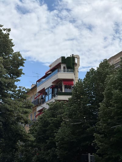 The image depicts a multi-story building in Nice, France, viewed from a low angle, possibly from the street level. The building features a modern architectural style with visible balconies, windows, and exterior decor including a green wall and red awnings. The sky is partly cloudy, and trees frame the building on both sides. An antenna is visible on the facade. The picture appears to be taken on a sunny day. The building's rooftop has mechanical equipment and vegetation, suggesting potential residential or commercial use.