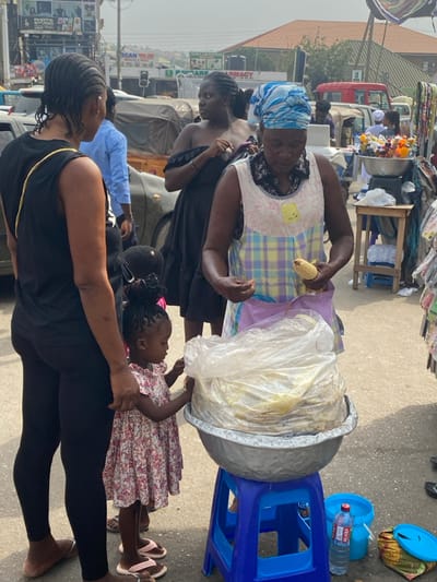 A woman in a patterned dress and headscarf holds corn, near a large bowl covered in plastic on a blue stool. A child in a floral dress reaches toward the bowl. Another woman, wearing black leggings and a top with a gold chain, stands nearby. The scene appears to be in a bustling street market, with buildings and vehicles visible in the background.