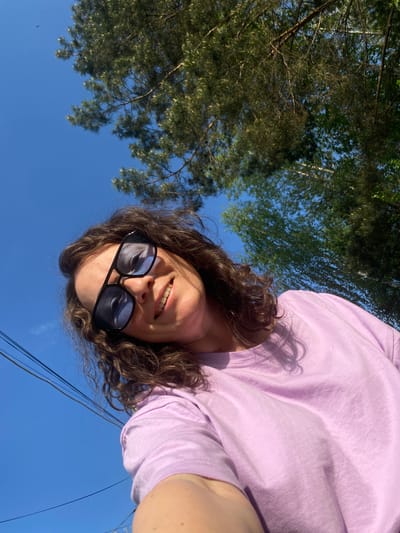 A woman with dark curly hair and sunglasses is taking a selfie in Ol'khovskoye, Russia, tilting the camera upward to capture the sky and trees in the background. She is wearing a light purple t-shirt. The sky is blue, and power lines are visible near the bottom of the frame. The trees are green and leafy.