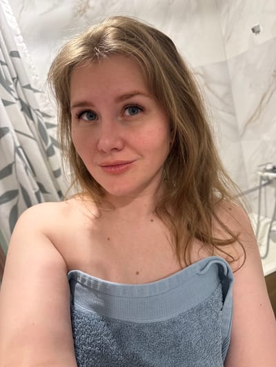 A young woman with light brown hair and blue eyes poses in a bathroom. She is partially covered by a light blue towel. The background includes a shower and a shower curtain with a gray and white leaf pattern. The location is likely a private residence. The city is Ферма, Russia.