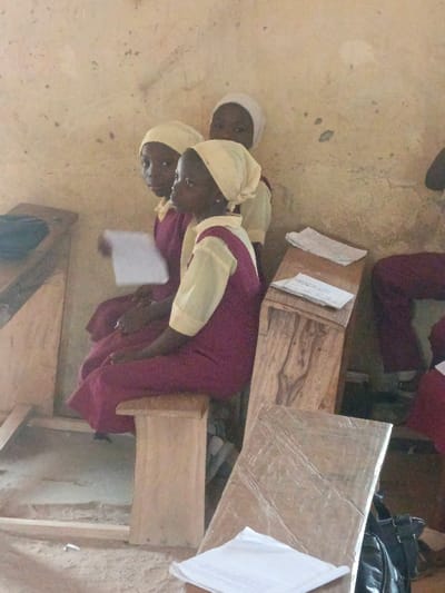 This image depicts three young females, identified as schoolgirls, seated inside a room in Mada, Nigeria. The girls are wearing uniforms consisting of maroon jumpers or dresses over light cream long-sleeved tops, with two wearing light cream head coverings and one wearing a white head covering. The foremost girl sits in a three-quarter profile facing right, while the girl behind her, also in uniform, holds a white sheet of paper in her left hand and looks towards the left. The third girl is partially visible behind them. They are seated on a basic wooden bench. The room contains rudimentary wooden desks or tables; one is partially visible on the far left, another in the mid-right foreground holds several folded papers, and a flat wooden surface with a folded white paper and a black bag rests in the immediate foreground. The walls are a light, distressed cream or light brown, showing discoloration and marks. The floor appears to be unpaved earth or sand.