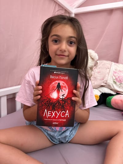 A young girl, with long brown hair and dark eyes, lies in bed, holding a book. The book's cover is predominantly red, featuring a silhouette of a woman against a large, pale moon, surrounded by bare, dark trees. The title, "ЛЕХУСА," is prominently displayed in white lettering. Smaller text on the book's spine indicates "Васил Попов," the author, and "storytel original." The girl is wearing a pink t-shirt and blue patterned shorts. She appears to be in a bedroom, with soft pink bedding and pillows behind her. The lighting suggests it could be daytime, possibly late afternoon. The girl's expression is pleasant, with a slight smile. A white crib frame is visible on the right side of the image. The overall scene conveys a sense of quiet relaxation and engagement with reading.