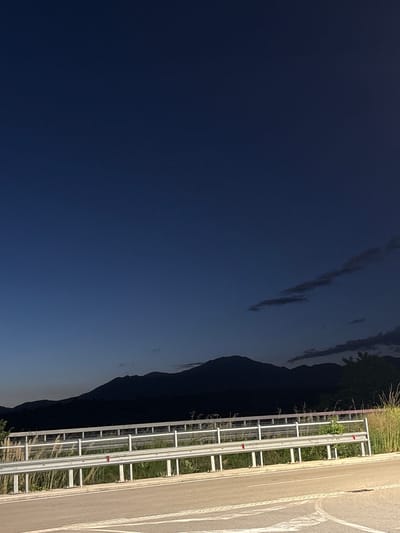 The image depicts a scene near Chuchuligovo, Bulgaria at dusk or night. To the left is a dark blue sky with scattered clouds. A silhouette of a mountain range extends along the right side of the sky. A roadside with a guardrail runs vertically on the right. The guardrail is white and metallic, with some vegetation growing around it. An asphalt road is visible to the right of the guardrail, marked with white lane lines.