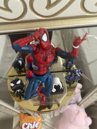 The image captures a close-up, eye-level view of an articulated Spider-Man action figure, centrally positioned and dynamically posed. The figure wears the classic red and blue suit with sculpted web patterns on the red sections. Its left arm is bent, with the hand near its head, while its right arm is extended, making the iconic "web-shooting" gesture with the thumb and pinky extended. The figure is supported by a clear plastic rod, which connects to a circular, highly reflective gold-colored base. A clear reflection of the figure's right hand is visible on this base.

Arranged on the same gold base are several smaller collectible items. To the lower left, there is a black, Pop! Vinyl-style keychain featuring the character Venom with an open mouth, fangs, and a prominent tongue. Another similar Venom keychain is partially visible behind the first. Behind the main Spider-Man figure, two small Lego-style minifigures, one standing and one partially obscured, appear to be Spider-Man in red and blue. To the right, a small, stylized purple figure, resembling Venom, stands on a matching purple disc base. A small white Lego minifigure headpiece or helmet on a tiny stand is visible near the main figure's left foot.

The background above the display base features a white surface with ornate gold scrollwork and a glittering gold band, suggesting a decorative piece of furniture or shelving. Below the gold base, blurred elements include an orange and white snack bag, partially visible with text resembling "Chic," and a soft, pink plush object to the bottom right. The lighting is bright and even, consistent with an indoor setting. No visual cues within the image indicate its location as the city of Ihtiman, Bulgaria.