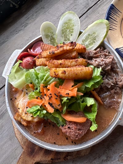 A close-up, overhead shot captures a sizzling platter of food served in a round, metallic dish with a wooden handle, placed on a weathered wooden surface. The dish contains a variety of ingredients: several thick-cut, golden-brown french fries are stacked in the center, nestled amongst crisp green lettuce leaves and shredded carrots. To the left, slices of red sausage and a small, pink patty are visible, partially submerged in a light brown sauce. The right side of the platter is filled with shredded meat, also in the sauce. Three oval-shaped slices of cucumber, their seeds clearly visible, adorn the top of the dish. Black sesame seeds are sprinkled over the carrots and lettuce, adding a touch of visual contrast. The overall presentation suggests a hearty and flavorful meal. The background reveals a rustic, wooden table, hinting at an outdoor or casual dining setting. There are no people present in the image. The provided location of Tây Hoa Lư Ward, Vietnam, is noted, but the image itself does not contain any specific visual cues to confirm or deny this precise location.