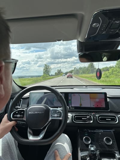 The image shows the interior of a car looking forward through the windshield. A person's left hand is visible on the steering wheel. The steering wheel has "SOUEAST" written on it. The dashboard features a large central touchscreen displaying a navigation map and other information. The car is driving on a paved road through a rural area with green fields and trees on either side. The sky is partly cloudy. In the distance, other cars are visible on the road. A decorative car air freshener, resembling a vinyl record, hangs from the rearview mirror. The car's interior appears modern and clean. The scene is likely occurring on a sunny day. The location is identified as Zav'yalovo, Russia.