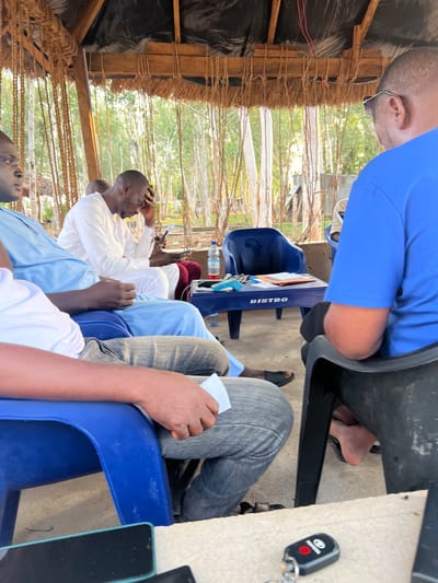 The image shows a group of people gathered outdoors, possibly for a meeting or discussion. The setting appears to be a rustic, open-sided shelter with a thatched roof. The subjects are several men seated on blue plastic chairs around a small, blue plastic table.
One man in the foreground, wearing a white long-sleeved shirt, is sitting with his hand on his forehead, appearing pensive or perhaps uncomfortable. Another man, wearing a bright blue shirt, is also visible, with part of his body obscured by a dark surface, possibly another chair or table. A person in grey pants is seated to the right. A Toyota car key is visible on a textured surface in the lower right quadrant.
On the blue table, there are several objects including a bottle of water, what appears to be a tablet or book, and a stapler or similar office supply. The word "BISTRO" is visible in white text on the side of the blue table.
The scene is lit by natural light, suggesting it is daytime. The background shows greenery and what looks like other structures, possibly indicative of a village or rural area. The overall mood seems informal, with the individuals engaged in their own thoughts or a collective activity. The location cues, such as the rustic shelter and the presence of the Toyota key, are consistent with the provided location context of Mayo Dasa, Nigeria.