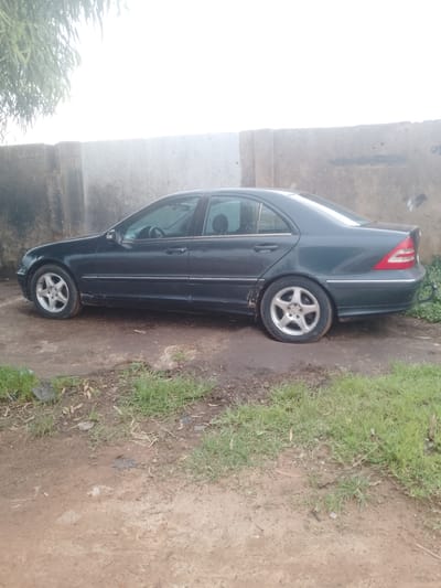 A dark green Mercedes-Benz sedan is parked on a dirt and grass area next to a concrete wall in Unguwar Saya, Nigeria. The car is facing left and appears to be in a state of disrepair, with some rust visible on the lower body panels. The area around the car is unpaved, with patches of green grass and loose dirt. The concrete wall behind the car is weathered and shows signs of graffiti. A tree with long, thin leaves is visible in the upper left corner of the image, casting a slight shadow. The sky is bright and overcast.