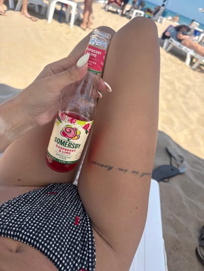 A person's legs are the focus of this close-up shot taken at a beach. The person is lying down on a beach chair on the sand, with other beachgoers and lounge chairs visible in the blurred background. The person is wearing a black and white checkered bikini with a cherry pattern. They are holding a bottle of Somersby Raspberry & Lime flavored cider. A tattoo is visible on their upper thigh. The scene suggests a sunny day at the beach, likely during the daytime given the brightness and beach setting. The overall mood appears relaxed and summery. The visible text on the bottle reads "SOMERSBY RASPBERRY & LIME FLAVOURED REFRESHING CIDER ALC. 4.5% VOL.".