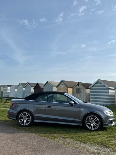 A gray Audi A3 convertible, with its black soft-top roof closed, is parked on a strip of green grass. The vehicle is oriented facing right, showcasing its multi-spoke alloy wheels. In the immediate foreground, a dark, narrow paved path runs parallel to the car. Behind the vehicle, a linear row of approximately ten wooden beach huts extends across the frame, receding towards the left. These huts display diverse paint schemes, including light blue, white, pale yellow, and weathered natural wood, with some featuring horizontal stripe patterns. A low wooden barrier or fence is visible between the car and the huts. The sky above is a clear, pale blue with scattered wispy white clouds. No individuals are present. This scene is located at Thorpe Bay, Southend-on-Sea, United Kingdom.