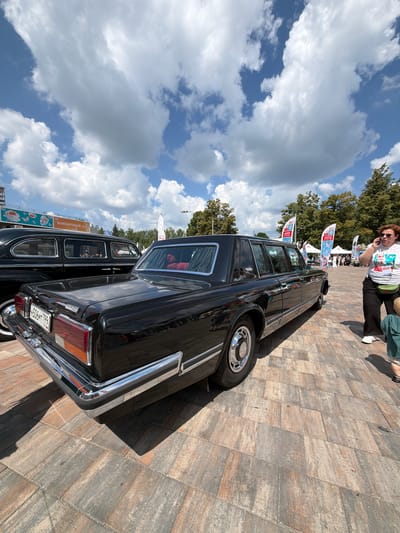 This is a medium shot, low-angle outdoor photograph taken on a sunny day with a partly cloudy sky. The main subject is the rear and side of a long, black, vintage limousine parked on a patterned stone pavement. The car has chrome accents on the bumper, along the bottom of the doors, and around the wheel wells. The license plate is visible and reads "H506MT 799". The interior of the car is visible through the windows, showing red seating.
To the left of the limousine, another black vintage car is partially visible. In the background, there are trees, tents, flags, and buildings. One banner visible on a building reads "КОНЦЕРТНЫЙ ЗАЛ" (Concert Hall).
On the right side of the image, a woman with curly red hair is standing and talking on a mobile phone. She is wearing a white t-shirt with a graphic print and dark pants. A green pouch is slung across her body. A small child in a green shirt is partially visible in the lower right corner of the frame. The overall setting appears to be an outdoor event or exhibition in a public space. The presence of the "Concert Hall" banner and the vintage cars suggests a possible cultural event or car show. Given the visual cues and context suggesting an event in Russia, and the specific mention of Moskovskiy, Russia, the scene is set in that location.