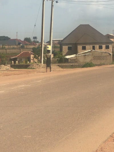 A woman is walking on a road in Kwang, Nigeria, carrying a yellow bucket on her head. She is wearing a patterned top and a dark skirt. In the background, there are several buildings, including a house with a brown tiled roof and a smaller structure with a corrugated metal roof. Power lines and utility poles are visible in the scene, suggesting an urban or semi-urban environment. The road appears to be unpaved, with dirt and gravel on the sides. The sky is clear and the lighting suggests it is daytime.