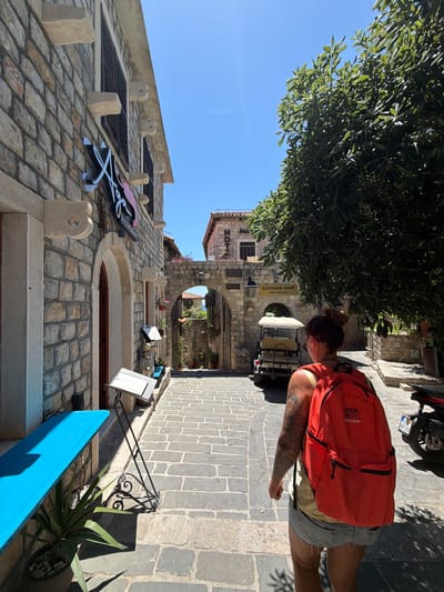 A woman with a red backpack walks down a narrow, stone-paved street in the Old Town of Ulcinj, Montenegro, on a sunny day. She is wearing denim shorts and a light-colored top, and her hair is pulled up in a bun. The street is lined with stone buildings, one of which has a sign for "HOTEL". A golf cart is parked on the side of the street, and a motorcycle is visible in the background. A large tree with green leaves provides shade on the right side of the image. The architecture of the buildings suggests a Mediterranean influence.