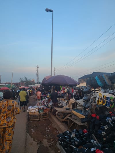 The image depicts a bustling street market in Nyanya, Nigeria, under a clear blue sky with a tall lamppost dominating the background. The market is characterized by numerous stalls selling a variety of goods, including footwear, clothing, and other assorted items.
In the foreground, a woman in a vibrantly patterned yellow and black outfit walks away from the camera, her back to the viewer. She carries a light-colored handbag. To her left, a narrow asphalt road is visible, with a dirt path bordering the market stalls on the right.
The market itself is a vibrant and crowded space. Stalls are set up along the path, covered by umbrellas and awnings. One prominent stall displays a large collection of footwear, including sandals and flip-flops in various colors and styles, hung on racks and piled on the ground. Other stalls showcase clothing and various trinkets.
Several individuals are seen browsing and interacting within the market. A man in a yellow t-shirt and shorts is walking past a stall, while another man in a striped shirt is visible further down the path. People appear to be engaged in shopping and moving through the market.
In the mid-ground, vehicles, including a car and motorcycles, are present, suggesting a busy thoroughfare connected to the market. The presence of tall telecommunication towers in the distance indicates an urban setting. The overall scene captures the lively atmosphere of a Nigerian street market, with goods, people, and infrastructure coexisting in a dynamic environment.