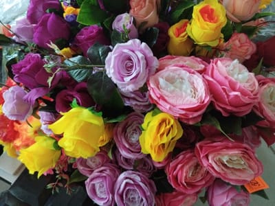 This is a close-up, high-angle, eye-level, indoor photograph of a vibrant bouquet of artificial flowers. The bouquet is predominantly composed of roses in various shades of pink, purple, and yellow, with some orange and red accents. The flowers are densely packed and fill the entire frame. The texture of the petals appears to be fabric-like, suggesting they are artificial. The lighting is bright, casting subtle shadows within the folds of the petals. There are no people or discernible actions taking place. The background is blurred and appears to be a neutral surface. No specific location in the city of Irpin, Ukraine can be identified from the visual information provided in the image.