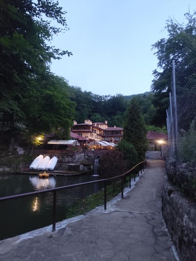 The image depicts a tranquil evening scene at a resort in Kostenets, Bulgaria. A body of water, likely a lake or pond, occupies the left side of the frame, with several white swan boats and a yellow paddle boat docked along its edge. Reflections of lights shimmer on the water's surface. A stone pathway, bordered by a metal railing, leads away from the viewer towards a multi-story building with a reddish-brown roof. The building appears to be a hotel or restaurant, with awnings extending over outdoor seating areas. Lush green trees and foliage surround the scene, with a hillside densely covered in trees in the background. A small waterfall cascades into the water on the right side of the building. The sky is a pale blue, suggesting dusk or early dawn. The overall atmosphere is serene and picturesque.