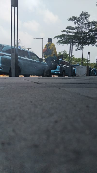 In a city street scene, a person wearing a yellow jacket and a backpack walks past several light blue cars parked in a row. The person is carrying a portable sprayer, suggesting they might be a gardener or involved in pest control. The backdrop features a clear sky with scattered clouds and trees, hinting at an outdoor, possibly public space in Bandung City, Indonesia. The low-angle perspective emphasizes the ground, which is paved with dark gray rectangular tiles.