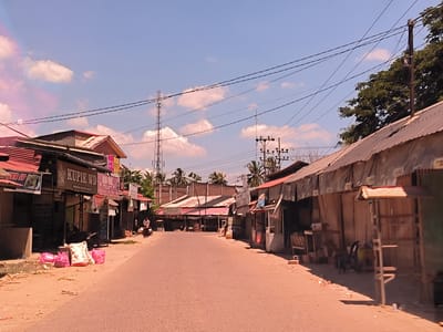 The image depicts a street in Keudepanteubreuh, Indonesia. The road, made of light brown asphalt, stretches into the distance, lined on both sides by single-story buildings. Most of the buildings appear to be shops or small businesses, some with corrugated metal roofs and makeshift awnings. Power lines crisscross overhead, supported by utility poles and a communication tower in the background. There are some pink bags of trash on the side of the road, with some individuals and motorcycles visible in the distance. The sky is mostly clear with scattered clouds.