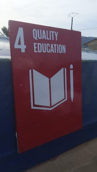 The image shows a red sign with white text and graphics, mounted on a blue surface. The sign reads "4 QUALITY EDUCATION" and depicts an open book with a pencil next to it. The sign is located in Limbe, Cameroon, at the coordinates 4.004 latitude and 9.211 longitude. The background behind the sign is blurred and out of focus, but it appears to be a building with a tiled roof and a wooden door. The sign is likely part of a larger display promoting quality education. The image captures the sign in a fixed position against the wall, conveying information about the importance of quality education.
