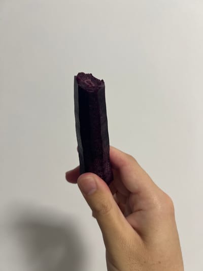 The image displays a close-up of a person's hand holding a dark purple, cylindrical object against a plain, light grey background. The object appears to be an edible item, possibly a piece of fruit, vegetable, or a snack bar, and has an irregular, somewhat textured surface. One end of the object shows a bite or break, revealing a reddish-purple interior with a fibrous or pulpy texture. The person's fingers are wrapped around the object, holding it horizontally across the frame. The lighting is bright and uniform, suggesting an indoor setting. No specific location cues, time of day, weather, or discernible emotions are evident from the image itself, apart from the provided context that it is from Mogán, Spain. There is no visible text.