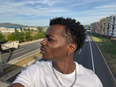 A dark-skinned male, likely a young adult, is depicted in a profile shot from the chest up, positioned on an elevated structure in Ripollet, Spain, overlooking a highway. He is facing towards the right side of the frame with a neutral expression, gazing off-camera. He has short, curly dark hair, a light mustache, and stubble. He wears a white ribbed crew-neck t-shirt, a silver link chain necklace, and a silver hoop earring in his visible left ear. The bright sun illuminates his face from the upper right, casting soft shadows.

The background reveals a multi-lane highway running horizontally below the subject, with two distinct carriageways separated by a grassy median. A white garbage truck travels on the left carriageway, moving left, while a white passenger car is visible on the right carriageway, also moving left. Dry grass and green vegetation line the highway. To the left, in the middle ground, are several light-colored industrial or commercial buildings against a backdrop of distant, verdant mountains under a partly cloudy blue sky. To the right, a long block of multi-story residential buildings stretches parallel to the highway. These buildings feature varied, faded pastel facades in shades of pink, yellow, blue, green, grey, and white, with numerous balconies displaying clotheslines and external air conditioning units. The scene is brightly lit by natural daylight.