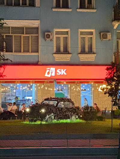 In Dushanbe, Tajikistan, at night, a cafe named "SK" is visible. The cafe has a bright red awning with "SK" logo. Inside, string lights illuminate the seating area. Outside, a vintage car covered in dark foliage is parked in front of the cafe, with its headlights on. Several people are seated at tables on the grass area in front of the cafe, enjoying the evening. The building behind the cafe is light blue with several windows. An air conditioning unit is mounted on the building's facade. The scene is set against a backdrop of a city street at night, with traffic lights and streetlights faintly visible in the distance.