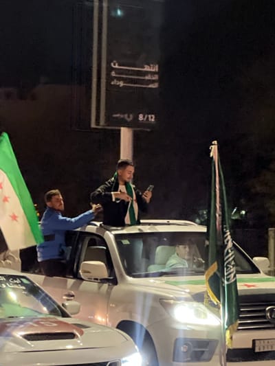 The image captures a nighttime scene, likely on a street or road in Al-Qanawat Municipality, Syria. Several men are engaged in what appears to be a celebratory or demonstrative event, leaning out of the windows of white vehicles, which resemble SUVs or vans.

In the center, a man with a beard, wearing a black jacket and a scarf patterned with the Syrian opposition flag (green, white, black stripes with three red stars), leans out of a vehicle, holding a mobile phone. Below him, another man in a blue shirt also leans out, holding a larger Syrian opposition flag that extends downwards. On the front of the vehicle they are leaning from, a prominent green flag with white Arabic calligraphy, likely depicting the Shahada ("La ilaha illallah, Muhammadun rasulullah"), is mounted. A small emblem resembling the Syrian opposition flag is also visible on the side of this vehicle near the headlight. Another white vehicle is partially visible in the foreground, showing a red square with the number "10" on its side.

The time of day is clearly night, as indicated by the dark background and the illuminated headlights of the vehicles. In the background, a dark street sign is visible with Arabic text. Parts of the text readable include "الله" (Allah), "أكبر" (Akbar), "وداد" (Widad), and the numbers "8/12." The overall atmosphere appears enthusiastic and active.