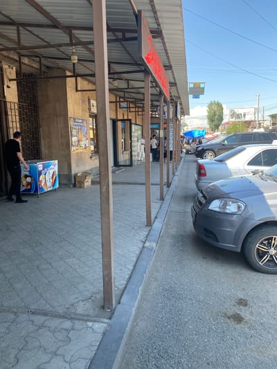 The image depicts an outdoor scene in the city of Gyumri, Armenia, likely a market or a commercial street. In the foreground on the left, a man in a black t-shirt and black pants is standing next to a colorful ice cream freezer. The freezer has an image of ice cream cones and scoops. Behind the man, a building with a covered walkway extends into the background. This walkway has several support poles and is lined with shops or stalls. A poster with Cyrillic text and images is affixed to the building wall.

To the right of the walkway, several cars are parked along the street. These include a silver sedan, a dark-colored SUV, and a gray Chevrolet. The street is paved, and there is a sidewalk area with patterned concrete slabs. The sky is visible above, with some clouds and power lines. The overall impression is a typical street scene in a commercial district, possibly during daytime hours given the lighting. The location is confirmed to be Gyumri, Armenia.