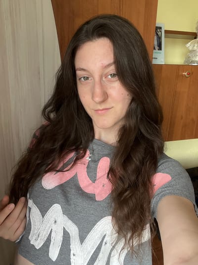 This is a close-up, portrait-style media file, oriented vertically, featuring a young person with long, dark, wavy hair. The subject's face is partially visible, with a soft smile and light-colored eyes. They are wearing a grey t-shirt with a large, abstract graphic in shades of pink and white. A delicate silver necklace with a small charm is visible on their chest.
The background is softly blurred, suggesting an indoor setting. To the left, a wooden surface is visible, possibly a shelf or a piece of furniture, with a small framed image resting on it. The image appears to be a photograph of two people. Some light-colored, possibly plastic packaging is also present on the shelf. The overall lighting suggests daytime, and the scene evokes a casual and relaxed mood. The visible text on the small framed image reads "e x i m a" and "c m a". The context of Popintsi, Bulgaria, provides a geographical reference for the location.
