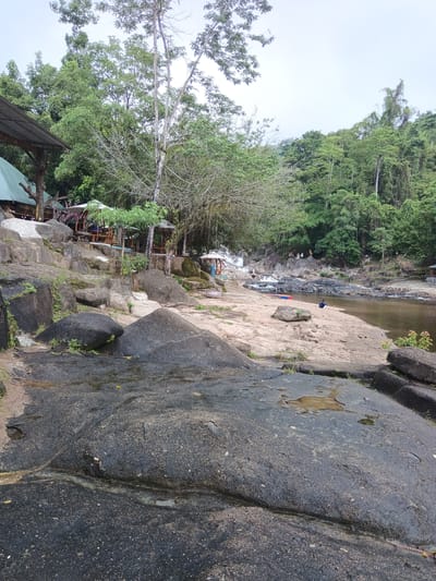 The image depicts a natural scene with a river and lush green foliage. In the foreground, large, dark, wet rocks are visible, some with shallow pools of water. A sandy bank leads to a river with a rocky, shallow flow. On the bank, a person is sitting, and a red and blue raft is placed on the sand. Further in the background, several people are gathered near a cascading waterfall and rocky outcrops. Several small structures with thatched roofs and open-air seating areas are scattered along the riverbank and on higher ground amidst the trees. The environment appears to be a park or recreational area within a tropical or subtropical region. The location is stated to be in the city of Khanh Vinh, Vietnam, suggesting a natural attraction within that area. The sky is overcast, contributing to a diffused light.