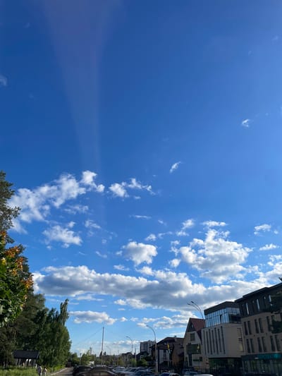 The image depicts a street scene in Brovary, Ukraine, under a bright blue sky with scattered white clouds. In the foreground, on the left, is a lush green tree with some autumn-colored leaves. To the right of the tree, a paved road is visible, lined with parked cars. Further down the road, several multi-story buildings are present, featuring modern architectural designs with large windows and balconies. One building has a sign with Cyrillic text. In the background, more trees and the outline of distant structures can be seen. The sky is a dominant feature, with sunbeams appearing to emanate from a point high above, creating a luminous effect through the clouds. Two individuals, appearing as pedestrians, are walking on a path in the lower left portion of the image. The overall impression is of an urban environment on a clear, sunny day.