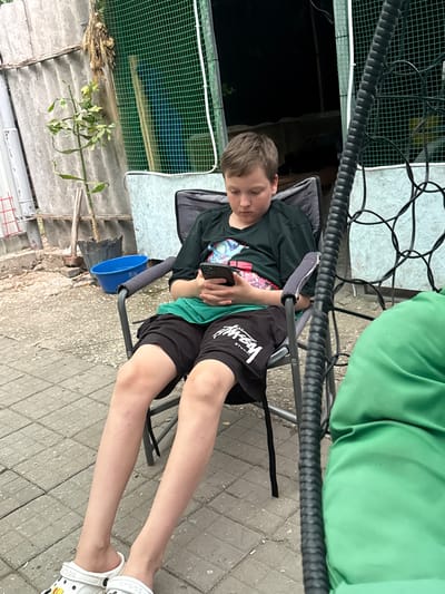 A young boy, appearing to be around 10-12 years old, is seated in a dark gray folding chair outdoors. He is wearing a black t-shirt with a colorful graphic design, a bright green undershirt, and black shorts with white lettering. The boy is looking down and holding a smartphone in his hands. His legs are extended forward, and he is wearing white Crocs-style sandals.
The setting appears to be a backyard or an outdoor area with a paved ground made of gray square tiles. To the left of the boy, there is a corrugated metal fence and a small potted plant with green leaves. A blue plastic basin is visible near the plant. Behind the boy, a green mesh netting is stretched on a frame. To the right, a portion of a green cushioned seating area is visible, suggesting it might be part of a swing or hanging chair. The overall environment suggests a casual outdoor space, possibly in a residential area. The location is identified as Советское сельское поселение, Ukraine.
