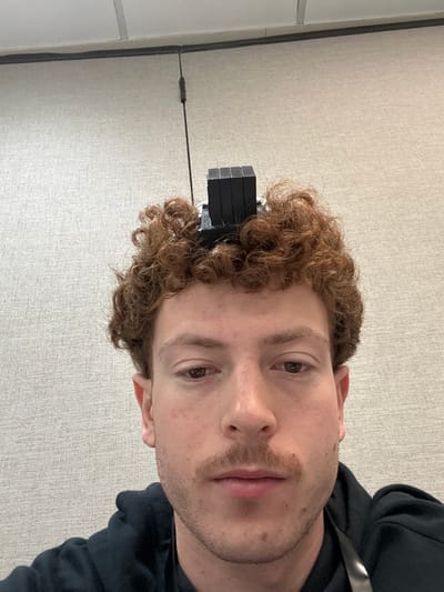 An image depicts a close-up, head-and-shoulders view of a young adult male, appearing Caucasian, positioned indoors. The subject has reddish-brown, curly hair and a mustache. His eyes are dark and gaze directly forward. A black tefillin shel rosh, a square box with a multi-segmented rectangular projection, is worn on the top of his head, partially obscured by his hair. He is wearing a dark-colored hooded garment, and a dark lanyard is visible over his right shoulder. The background consists of a light-colored, textured wall. Above the wall, a section of a suspended acoustic ceiling with white tiles and dark grid lines is visible. A thin, dark cable or cord is present vertically against the upper portion of the wall. No specific location within Fairfield, United States of America, is identifiable from the visible details.