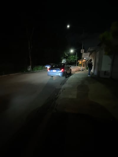 A blue sedan is parked on the side of a dark, paved road at night. The car's taillights are illuminated. A person, wearing a dark jacket and jeans, stands on the sidewalk next to a building. The building appears to be a storefront with a sign. Streetlights illuminate parts of the scene, casting long shadows. The environment suggests a residential or commercial street in Huixquilucan de Degollado, Mexico, given the presence of a building with a Spanish sign and a common vehicle model. The overall lighting conditions indicate nighttime.
