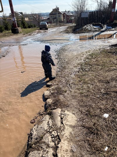 A person stands in a large puddle of muddy water on a street in Ферма, Russia. The water covers most of the road, reflecting the sunlight. To the left, a car is partially visible driving through another section of the flooded street. Buildings and trees line the road in the background. The person is wearing dark clothing and a hat and appears to be gesturing with one hand. The edges of the flooded area are bordered by dirt, gravel, and some vegetation.