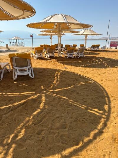 The image depicts a beach scene at the Dead Sea in Tamar Regional Council, Israel. The foreground is dominated by golden sand, casting long shadows from large, yellow-topped umbrellas. Several empty lounge chairs with yellow covers are arranged on the sand, suggesting a quiet moment on the beach. In the middle ground, the calm, turquoise waters of the Dead Sea are visible, with several people in the water. A small, open-sided shelter is situated near the water's edge, providing shade. In the background, hills or mountains can be seen on the horizon under a clear, bright blue sky. A sign with Hebrew writing is posted on a pole near the umbrellas, likely indicating beach regulations. The overall impression is one of a serene and inviting beach environment.