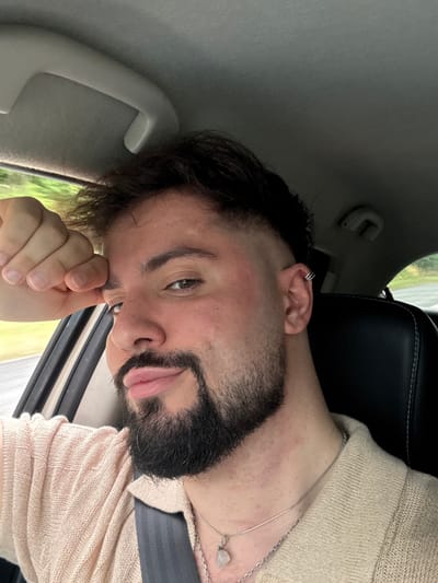 A bearded man in a car, wearing a seatbelt and a light-colored shirt, gazes into the camera. He has dark hair, multiple earrings on one ear, and a silver necklace with a clear stone pendant. His hand is near his face, possibly touching his nose. The car interior is visible, with a black headrest and a gray ceiling. The background appears to be a glimpse of greenery through the car window.
