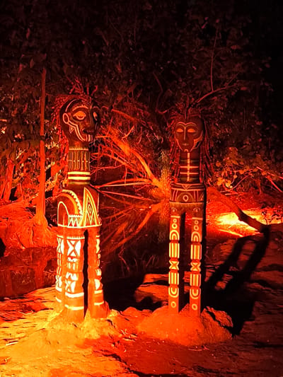 The image depicts two tribal statues illuminated by warm, orange lighting, set against a backdrop of dark foliage and trees. The statues, one on the left and one on the right, are carved from wood and adorned with intricate white patterns. The statue on the left has a broader torso and legs, while the statue on the right is more slender. Both statues have distinct facial features and headdresses. The ground around the statues appears to be rocky and uneven, with shadows cast by the statues and surrounding vegetation. The overall atmosphere is mystical and evocative, suggesting a ritualistic or ceremonial setting. The location of this scene is the Taman Sekartaji park in Kediri, East Java, Indonesia.
