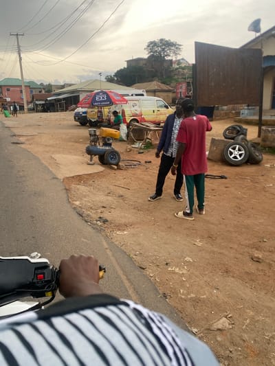 The image depicts an outdoor scene in Kabong, Nigeria, possibly a roadside market or workshop. In the foreground, a hand is visible operating a motorcycle's handlebar, suggesting the photo is taken from the perspective of a motorcyclist. The middle ground shows three individuals, two men and one woman. One man is wearing a dark blue jacket and a patterned shirt, while another is wearing a red t-shirt and green and white striped pants. The woman is wearing a green t-shirt and is seated. They appear to be interacting or observing something.
Adjacent to them is a yellow and white van with branding that includes "DANGOTE 3X LASTS LONGER YIELDS MORE." Nearby, there is a blue air compressor with a yellow tank and wheels. Several tires, including one with a silver rim, are stacked on concrete blocks to the right of the frame. The ground is unpaved, composed of reddish-brown dirt and some scattered debris. In the background, buildings with corrugated metal roofs and residential structures are visible, along with a utility pole with numerous overhead wires. The sky is overcast. The overall impression is a bustling, somewhat cluttered environment typical of a commercial area in Nigeria.