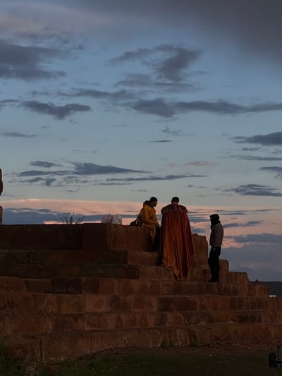 The image shows three people standing on a large stone structure resembling stairs or terraces. The sky in the background is filled with clouds, with a blend of blues, grays, and a hint of pink near the horizon. The stone structure appears to be old, with a weathered and uneven surface.
From the bottom of the structure to the top, one person is sitting down while the other is standing up wearing a long orange robe. Lastly, there's another person wearing darker clothing standing alone on a higher level. Vegetation is visible on the ground to the right of the structure. There is an object behind the person on the top step, possibly a camera on a tripod.
Given the reference to Anipemza, Armenia, this could be a part of the archaeological site of Ani, a ruined medieval Armenian city known for its stone architecture. However, without further context, it is impossible to confirm this definitively.