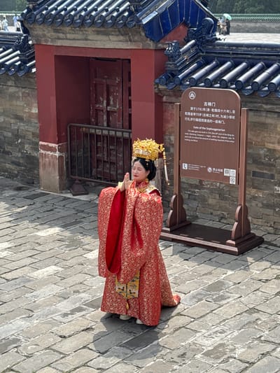 The image shows a woman in traditional Chinese dress, kneeling in what appears to be a courtyard in Dongcheng District, China. She wears an elaborate golden headdress and a red patterned robe. Her hands are clasped together in front of her. Behind her is a red building structure with a dark blue tiled roof. To the side of the woman is a sign in both Chinese and English, marking the location as the "Gate of the Septuagenarian," which was built in 1781 to reduce the amount of walking required for Emperor Qianlong. The courtyard is paved with grey stone bricks.