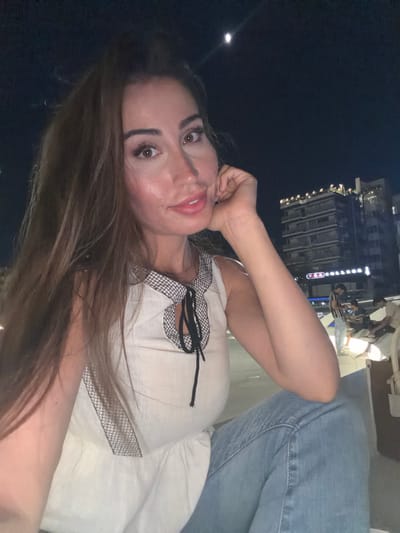 A woman with long, brown hair and full lips, wearing a white sleeveless top with black trim and blue jeans, poses for a selfie at night in Nicosia, Cyprus. She rests her chin on her hand, looking directly at the camera. In the background, a building with illuminated windows is visible, along with a partial view of the moon in the dark sky. Several people are blurred in the background, some seated. The environment appears to be an outdoor urban setting.
