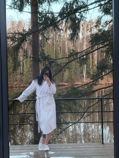 A full-length image captures a person's reflection in a large window overlooking a forest. The person, with long dark hair, is wearing a white bathrobe and white slippers, holding a smartphone to take a picture, which partially obscures their face. The reflection makes it appear as though the individual is standing amidst the trees.
The scene outside the window is a dense forest, composed of both coniferous trees and bare deciduous trees, possibly birches, identifiable by their light-colored trunks. The absence of leaves on many trees suggests an autumn, winter, or early spring season. The light appears soft, indicating daytime, possibly overcast or late afternoon. A dark window frame borders the view, and a section of a light-colored wooden structure is visible to the right, also reflecting in the glass. No text is visible in the image. The setting evokes a serene, natural environment, consistent with locations like Мишкино, Russia.