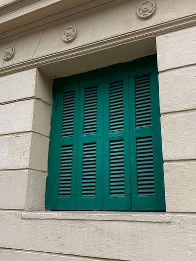 An upward-angled shot captures a recessed, dark green louvered structure framed by a light-colored architectural facade. The central subject is a rectangular panel, possibly a shutter or a vent, composed of four distinct horizontal sections, each filled with vertical louvers. The green surface appears to be painted wood or metal, showing some subtle wear and hinge details between the sections, suggesting it might be foldable or operational.
Surrounding this green element is a robust, light beige or off-white structure, likely made of concrete or stucco. This frame features prominent linear moldings and carved decorative rosettes with floral or star-like patterns, visible along the left vertical beam and partially on the upper and lower sections. The surrounding material has a textured, slightly rough finish, contrasting with the smoother appearance of the louvered panel.
The perspective is looking up, with light from above casting subtle shadows that highlight the three-dimensional nature of the architectural details and the depth of the recessed green panel. There are no people or specific activities depicted. The scene is static, focusing on architectural details. The overall impression is one of a functional and decorative architectural element, possibly from an older building in Marousi, Greece, given the provided context. There is no visible text in the image.