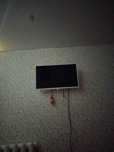 The image shows a television mounted on a wall with floral wallpaper. A red object, possibly a remote control or a signal receiver, is attached to the wall below the TV. A black cable runs down from the TV. In the lower left corner, part of a white radiator is visible. The ceiling above the wall has a circular vent. The image appears to be an interior shot, likely a domestic setting in Orsk, Russia. The lighting is dim.