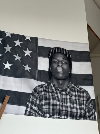 A black and white photograph depicts A$AP Rocky standing in front of an American flag. He is wearing a plaid shirt and a patterned baseball cap, with smoke coming from his mouth. The photograph is displayed on a wall in a room. The room appears to be modern, with white walls and a wooden accent piece. The photograph is a large print, taking up a significant portion of the wall. The overall impression is one of casual artistry.